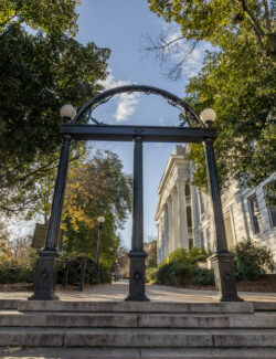 A black iron archway stands at the entrance to a university campus, surrounded by trees and historic white buildings under a clear sky.