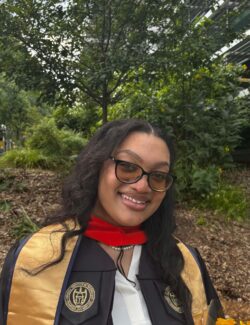 Pierre-Mitchell Photo A woman in graduation attire, wearing glasses and holding a bouquet of yellow flowers, stands outside near greenery and a modern building.